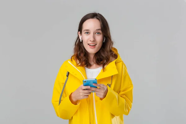 Mujer morena sonriente en un impermeable amarillo está leyendo notificación en el teléfono inteligente. El modelo usa auriculares inalámbricos. Concepto de comunicación . —  Fotos de Stock