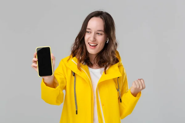 Mujer morena sonriente en un impermeable amarillo expresando emoción de éxito mostrando la pantalla del teléfono inteligente aislado sobre fondo gris. Concepto de éxito . — Foto de Stock