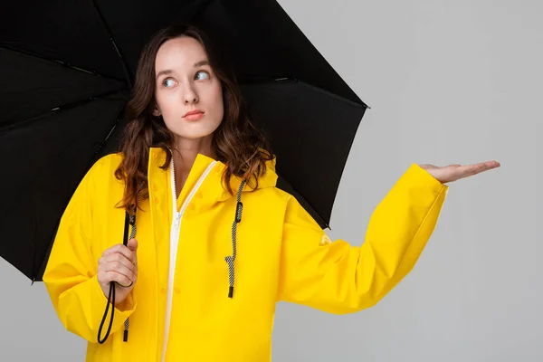 Linda chica morena escondida bajo un paraguas negro vestida con impermeable amarillo. Modelo de comprobación de si la lluvia ha comenzado. Prepárate para el mal tiempo . —  Fotos de Stock