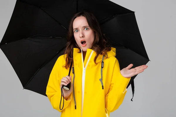 Pretty brunette girl hiding under black umbrella dressed into yellow raincoat. Model checking if rain has started. Get ready for bad weather.