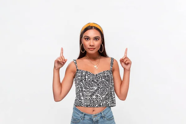 Pretty african-american teenager in a headband, sleeveless t-shirt and jeans pointing up standing isolated over white background. Copy space of advertising. — Stock Photo, Image