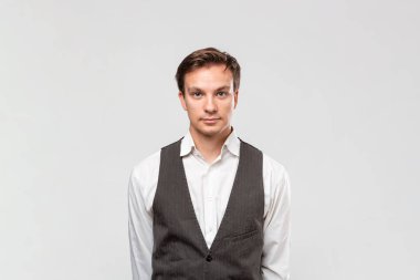 Beyaz gömlekli, gri yelekli, kapalı yakışıklı bir adam gri arka planda tek başına duran kameraya bakıyor..