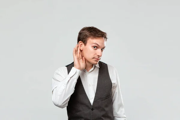 Jovem bonito com uma camisa branca e colete cinza segurando a mão perto do ouvido ouvindo algum boato interessante . — Fotografia de Stock