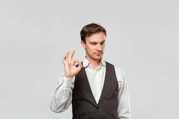Homem bonito em uma camisa branca e colete cinza mostrando OK sinal isolado sobre fundo cinza claro . — Fotografia de Stock