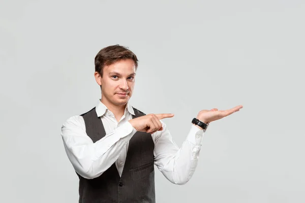 Hombre guapo en una camisa blanca y chaleco gris sosteniendo la palma de una mano para la colocación del producto o la publicidad . — Foto de Stock