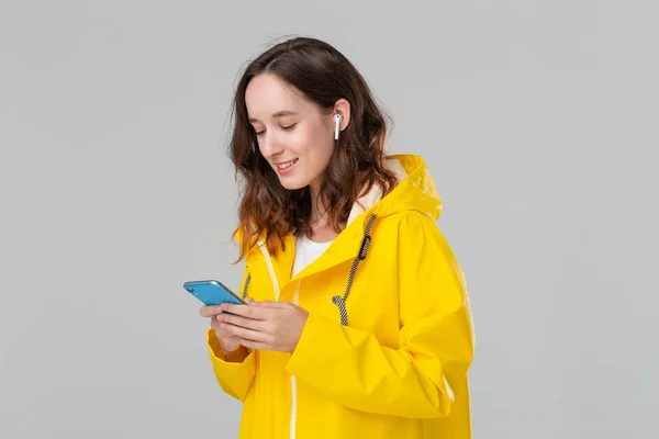 Mujer morena bonita en un impermeable amarillo está leyendo notificación en el teléfono inteligente aislado sobre fondo gris. Modelo lleva auriculares inalámbricos — Foto de Stock
