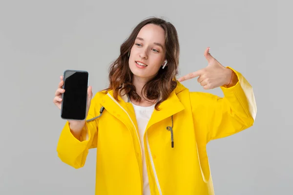 Mujer morena excitada en un impermeable amarillo que muestra la pantalla de un teléfono móvil aislado sobre fondo gris. Modelo lleva auriculares inalámbricos . — Foto de Stock