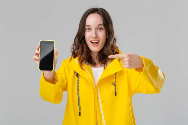 Mujer morena excitada en un impermeable amarillo que muestra la pantalla de un teléfono móvil mirando a la cámara aislada sobre un fondo gris. Modelo lleva auriculares inalámbricos . — Foto de Stock