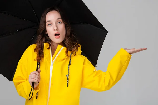 Chica morena sorprendida escondida bajo paraguas negro vestido con impermeable amarillo. Modelo de comprobación de si la lluvia ha comenzado . —  Fotos de Stock