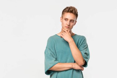 Yeşil tişörtlü düşünceli genç adam gri arka planda izole edilmiş çenesinin yanında el ele tutuşuyor..