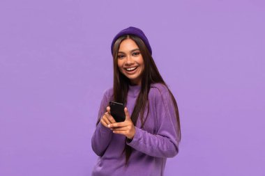 Smiling african-american girl in a hat and sweater holding smartphone isolated over purple background. clipart