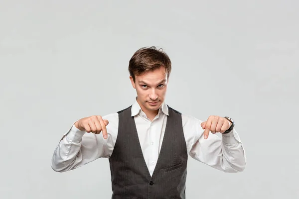 Homem bonito com uma camisa branca e colete cinza mostrando com os dedos para baixo no espaço vazio para publicidade . — Fotografia de Stock
