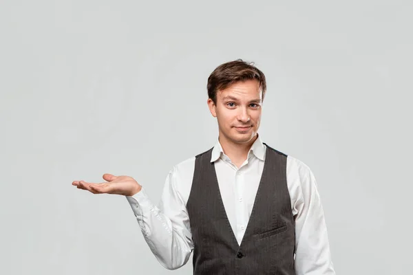 Hombre guapo en una camisa blanca y chaleco gris sosteniendo la palma de una mano para la colocación del producto o la publicidad . — Foto de Stock