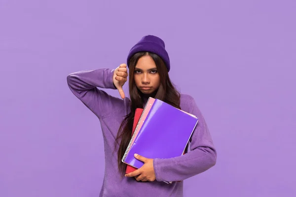 Triste adolescente afroamericano en un sombrero y suéter sosteniendo carpetas y mostrando el pulgar hacia abajo de pie aislado sobre el fondo púrpura . — Foto de Stock