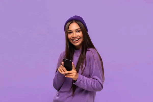 Sonriente chica afroamericana en un sombrero y suéter sosteniendo teléfono inteligente aislado sobre fondo púrpura . —  Fotos de Stock