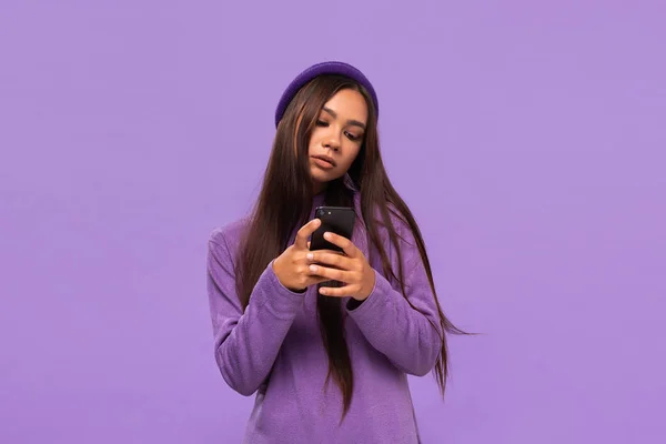 Chica afroamericana en un sombrero y suéter sosteniendo teléfono inteligente aislado sobre fondo púrpura . — Foto de Stock