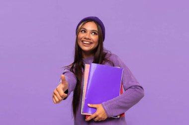 Şapka ve süveter giymiş gülümseyen Afro-Amerikan genç öğrenci elinde klasörler ve mor arka planda izole bir şekilde ayakta duruyor..