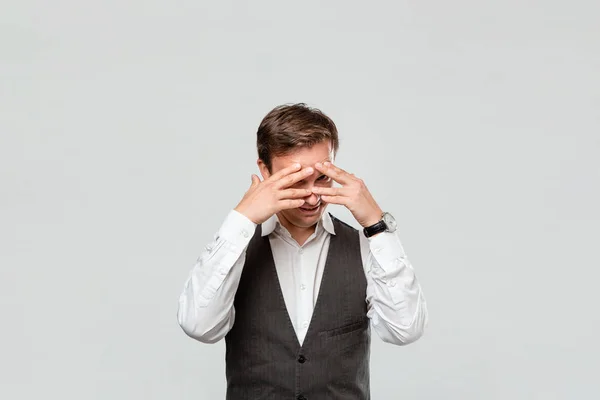 Hombre guapo con camisa blanca y chaleco gris escondido detrás de sus manos aislado sobre fondo gris . — Foto de Stock