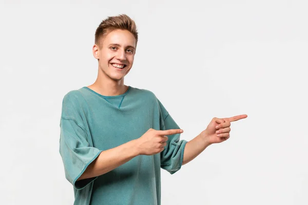 Przystojny młody człowiek w zielonej koszulce pokazuje palcami na pustej przestrzeni do reklamy po prawej stronie białego tła. — Zdjęcie stockowe