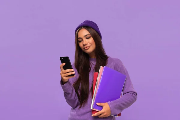 Pretty african-american teenager student in a hat and sweater holding folders and looking surprised checking notification on a smartphone standing isolated over purple background. — Stock Photo, Image