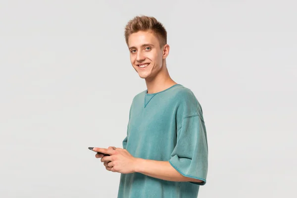Улыбающийся блондин-подросток в зеленой футболке держит смартфон и смотрит на камеру, изолированную на светло-сером фоне . — стоковое фото