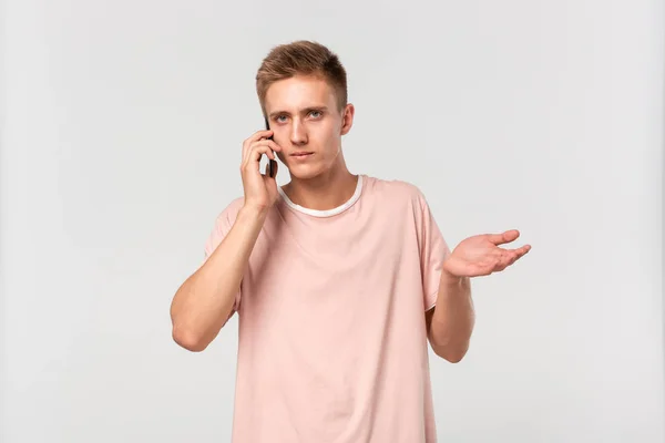 Adolescente rubia confusa en una camiseta rosa hablando en el teléfono inteligente, encogiéndose de hombros y mirando a la cámara aislada sobre un fondo gris claro . —  Fotos de Stock