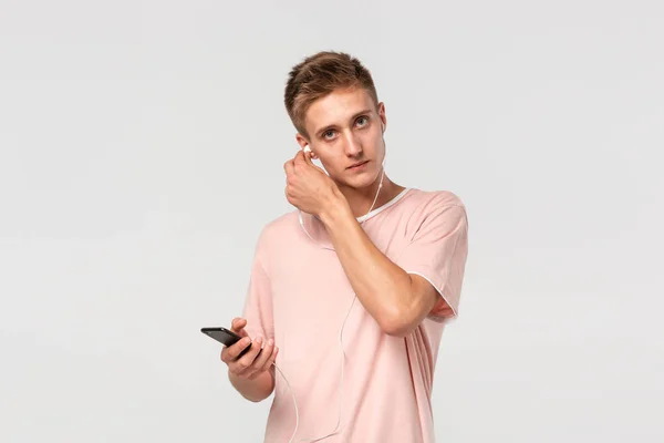 Adolescente bastante rubia en una camiseta rosa escuchando música en un teléfono inteligente con auriculares con cable y mirando a la cámara aislada sobre fondo gris claro . —  Fotos de Stock