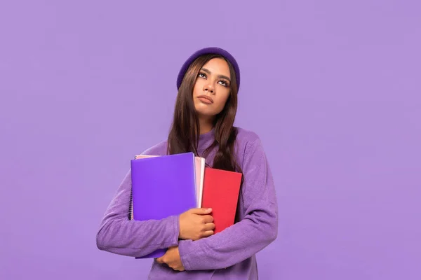 Adolescente afro-americano perturbado em um chapéu e camisola segurando pastas de pé isolado sobre fundo roxo . — Fotografia de Stock