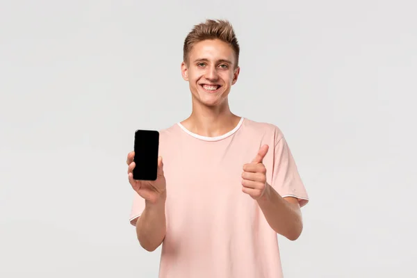 穿着粉色T恤的金发少女微笑着，指着被淡灰色背景隔离的智能手机屏幕. — 图库照片
