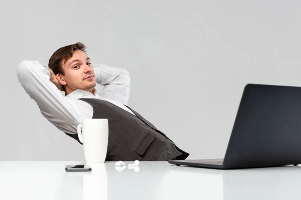 ホワイトのTシャツとグレーのベストのオフィスワーカーは、ノートパソコン、スマートフォン、ホットドリンクのカップと白いテーブルに座って休んでいます。. — ストック写真