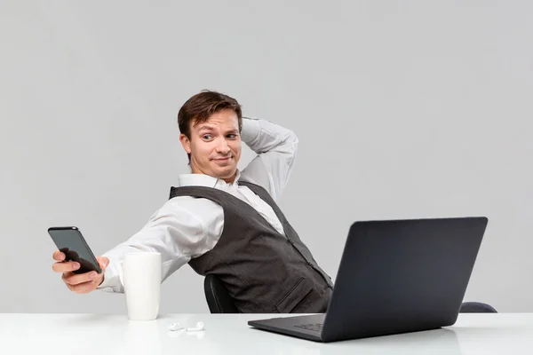 Empresário em uma camiseta branca e colete cinza verificando smartphone e bebendo café sentado na mesa branca com laptop . — Fotografia de Stock