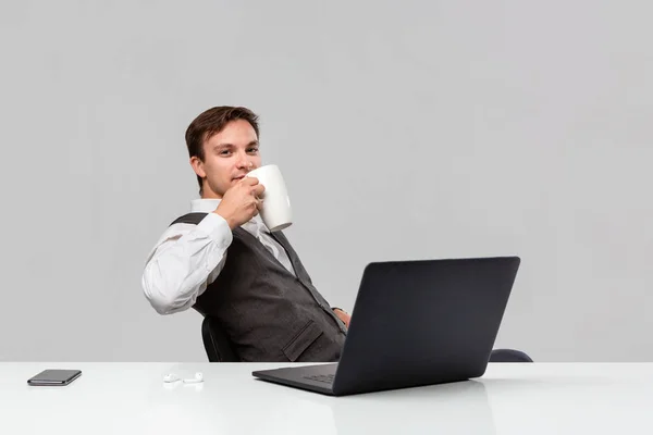 Empresario en camiseta blanca y chaleco gris bebiendo café sentado en la mesa blanca con portátil y smartphone . — Foto de Stock