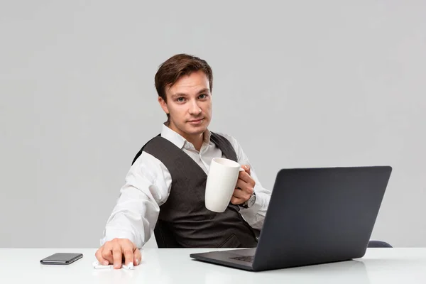 Empresario en camiseta blanca y chaleco gris bebiendo café sentado en la mesa blanca con portátil y smartphone . — Foto de Stock