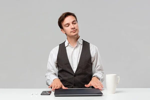 Bonito trabalhador de escritório em uma camiseta branca e colete cinza colocando as coisas em ordem em sua área de trabalho. Laptop, smartphone e xícara de café em uma mesa branca . — Fotografia de Stock