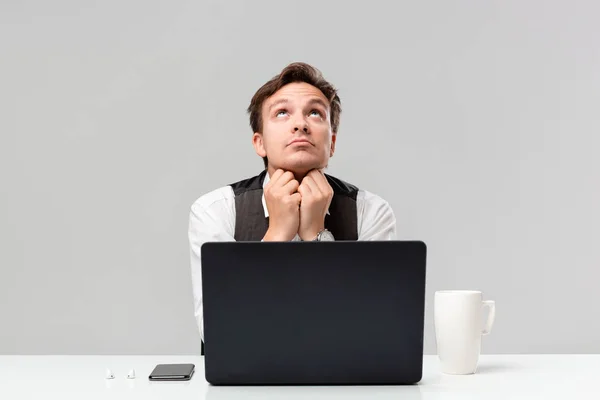 Homem bonito em uma camiseta branca e colete cinza expressando emoção de suplicar sentado na mesa branca com um laptop, smartphone e xícara de café . — Fotografia de Stock