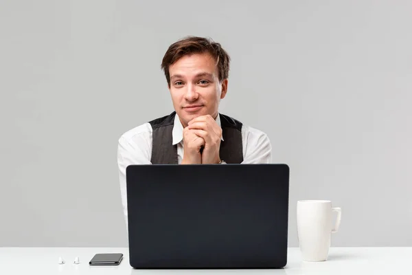 Empresário em uma camiseta branca e colete cinza trabalhando no laptop no escritório sentado na mesa branca com laptop e smartphone . — Fotografia de Stock