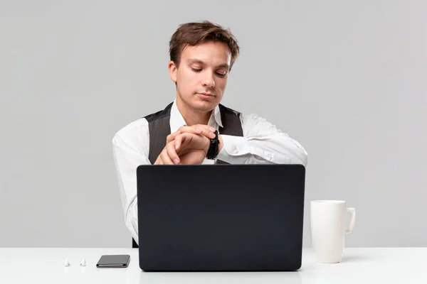 Bonito homem em uma camiseta branca e colete cinza verificando o tempo para consulta de negócios sentado na mesa branca com laptop e smartphone . — Fotografia de Stock