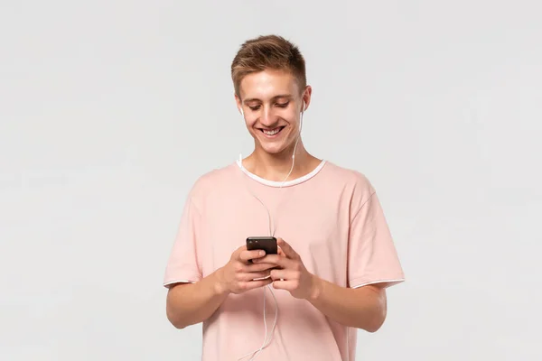 Adolescente rubia sonriente en una camiseta rosa escuchando música en un teléfono inteligente con auriculares con cable aislados sobre fondo gris claro . —  Fotos de Stock