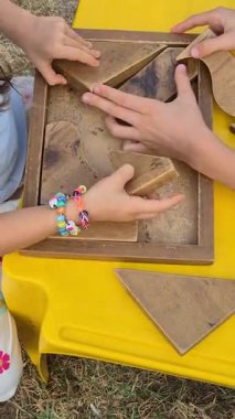 Children assembling a wooden puzzle game on a yellow table. Hands work together to solve and fit pieces, fostering concentration, teamwork, and playful learning. No faces visible.