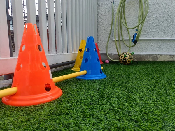 Turuncu koni sarı koni eğitim direği tatbikatı. Köşesinde hortum ve top olan yapay çimenler üzerinde. Açık hava sporları sahnesi yaratıyor.
