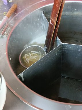 Metal çorba istasyonu, ahşap sapları ve fasulye filizi tutan buharlı et suyunda garnitür, sıcak havayla dolu mutfak sahnesi.