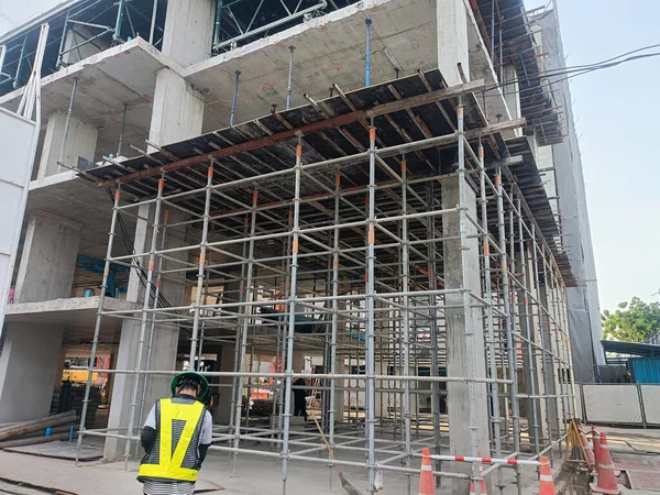 Çelik yelek giyen işçiler inşaat halindeki beton binanın çelik iskelesini, kentsel alan atmosferini alet ve dikkat konileriyle inceliyor.