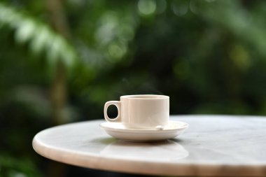 Açık hava bahçesindeki mermer masanın üstünde beyaz kahve fincanı, sakin sabah atmosferi, sığ alan derinliği, doğal ışık.