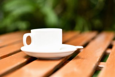 Beyaz kahve fincanı ve çay tabağı açık hava bahçesindeki ahşap masada kaşık, sıcak sabah ışığı ve sakin bir ruh hali.