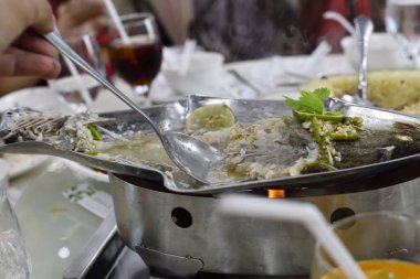 Masa başı ızgarasında baharatlı et suyuna buharlı balık, kaşık servisi, bitki süslemesi ve ortak yemek atmosferi.