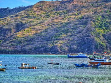 Lombok, Endonezya 5 Ağustos 2024. Huzurlu tekneler sakin bir turkuaz körfezde yavaşça sallanır yemyeşil bir tepe arkaplanı ile rahatlama amaçlı bir seyahat posteri veya masaüstü arka planı için mükemmeldir..