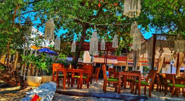 Lombok, Endonezya 5 Ağustos 2024. Gili Trawangan. Kumlu bir sahildeki açık hava restoranını davet etmek ahşap masalar, makrome fenerler ve bereketli ağaç örtüsü seyahat ve etkinlik promosyonları için harika..