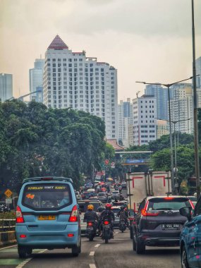 Jakarta, Endonezya 12 Nisan 2025. Binalar ve ağaçlar arasında yoğun trafik Jakarta caddelerinde yavaş ilerliyor; işe gidiş gelişleri ve kentsel yaşam zorluklarını göstermek için ideal.
