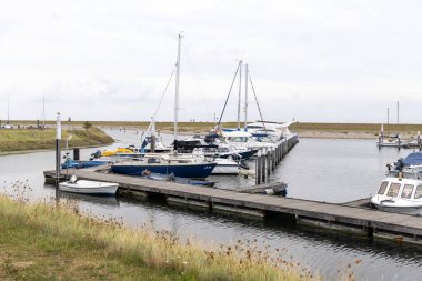 Ahşap rıhtımlara demirlemiş teknelerin olduğu liman sahnesi, çimenli alanlar ve sakin bir gökyüzü, su bazlı dinlenme için huzurlu bir ortam sağlıyor.
