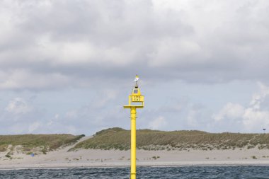 Canlı sarı renkli seyir şamandırası sakin sularda, arka planda kumlu kumullar ve çimenli kumullarla, teknelerin güvenli bir şekilde yol göstermesi için yerleştirildi.